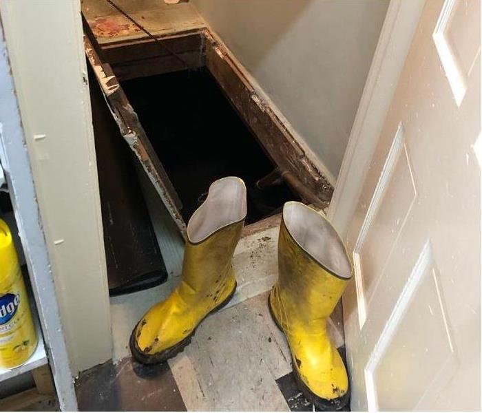 Open access door in the floor of a room with dirty yellow work boots sitting on the side.